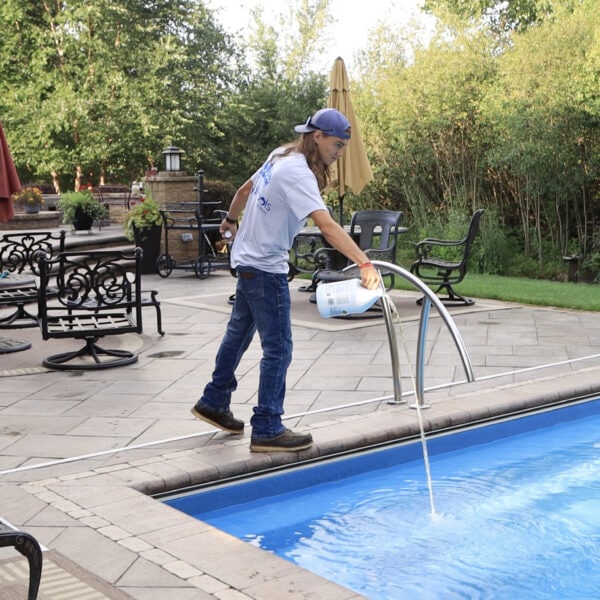 Photo of a man standing over the shallow end of a light-blue inground pool pouring a yellow liquid into the pool. Behind the pool is a stamped concrete patio with chairs and a retaining wall. Trees can be seen behind the patio.
